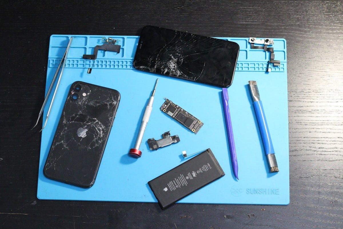 Disassembled broken phone with battery, screen, and repair tools on a blue mat.